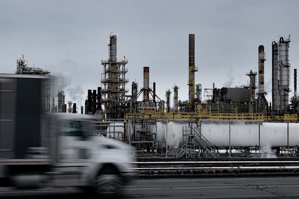A truck drives past a large oil refinery under a cloudy sky, capturing industrial transportation.